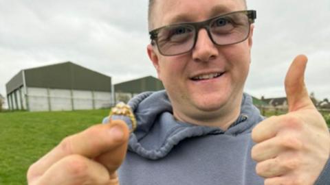 A man in his early 40s smiles as he stands in a field on a cloudy day. In one hand, he is holding up a gold diamond ring, while he gives a thumbs up with the other hand. He is wearing a grey hoodie and black-rimmed glasses, the lens of which are slightly tinted. There are large metal barns behind him.