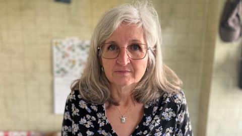 A woman with grey hair, glasses and a dark blue blouse with flowers is looking into the camera. She is standing in her kitchen. 