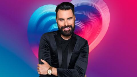Rylan Clark, wearing a black suit and standing in front of a pink and blue Eurovision branding