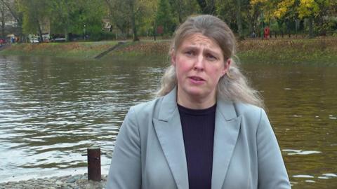 Rachael Maskell has a serious expression as she stands by the River Ouse, a wide waterway in central York. She has long blond hair, tied back and wears a pale green jacked and black T-shirt. Behind her, steep grassy banks, topped by green trees, lead down the the river. 