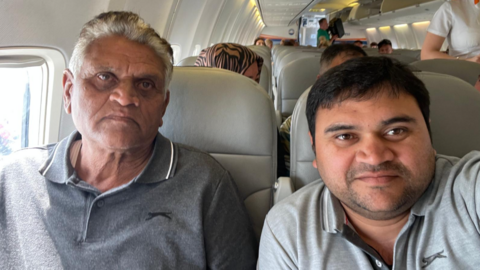 Umang and Kirat Patel take a selfie on board a plane. Both men are wearing grey polo shirts. Umang Patel is smiling slightly while his father is looking quite serious. The seats of the plane are light grey. People can be seen sitting behind Umang and his father and a stewardess is walking up the aisle.
