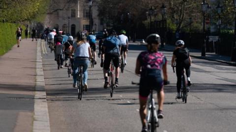 Members of the public cycling round Regent's Park, London. Cyclists in helmets travel along a stretch of road on a warm looking day. 