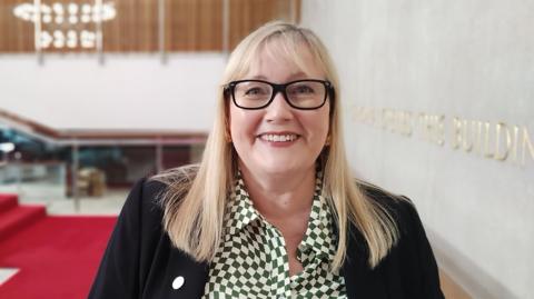 Karen Kilgour smiling at the camera. She has medium-length straight blonde hair with a fringe. She is wearing black-rimmed glasses, a green-and-white chequered-patterned shirt and a black suit jacket.