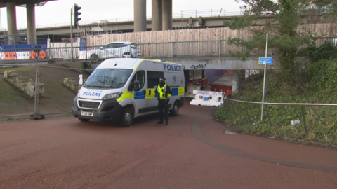 A police van and police officer by an underpass where a woman was attacked and raped. The underpass is sealed off, while cars are driving by on the road above it