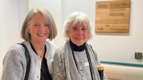 Nic and Dianne in a mid-shot, smiling to camera, both wearing grey tops and standing in front of a wooden plaque which says 'The Vanessa Kyte Gamma Suite' on the wall of a hospital corridor