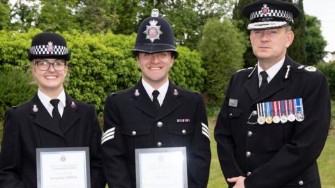 PC Sam Hilbery and Sgt Alex Farr with Essex Chief Constable Ben-Julian Harrington, all in police uniform, facing the camera, they all have police hats on. Sam and Alex are holding certificates in a frame. 