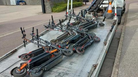 Rows of e-scooters on the back of a low-loader lorry which is parked on a road next to a pavement.
