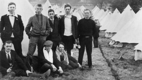 Black and white image of eight men looking to camera with lots of tents behind them