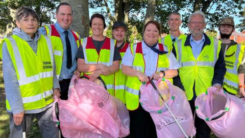 A group of eight men and women wearing high-visibility vests and carrying pink litter bags and long litter-picking grabbers. They are standing in a park in front of trees.