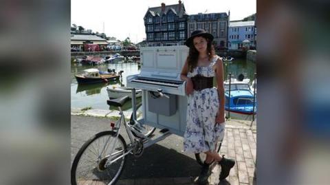 Chloe Marie Aston, in a blue and white dress, stands next to her bike, which has a piano attached to the front of it