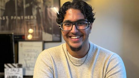 Zane Alsaroori, a young man from Yemeni heritage, with glasses, middle-parted curls, and a short goatee.