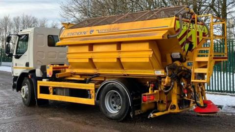 A gritting truck, with a white cab and yellow trailer containing grit, drives along a road with snow covering the pavement.