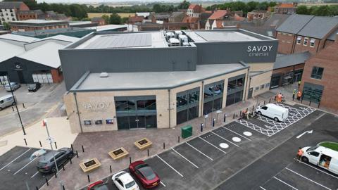 Aerial view of the new Savoy Cinema  a three-storey building in light stone with large grey windows and a grey cladded roof with the cinema's name in silver lettering. There is a car park at the front and a row of shops to the right.