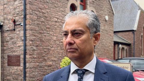 Obi Hassan is a man with grey hair. He is wearing a grey suit with a white shirt and navy tie with a circular pattern on it. He is staring to the left side of the photo. The background is a stone community centre.