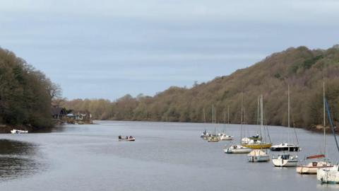 A lake with trees either side and a number of boats lined up on the right and another in the centre of the picture. A house can be seen on the lake's shore in the distance as the lake bends around a corner to the left.