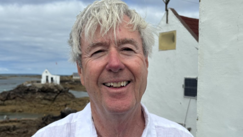 Richard Miles has grey hair. He's wearing a white collared linen shirt, standing beside a white-painted building on a rocky coastline. In the background, shallow coastal waters surround dark rocks, and a small white building sits on a rocky outcrop near the shore. The sky is overcast with layers of grey clouds, and the scene is lit by natural daylight.