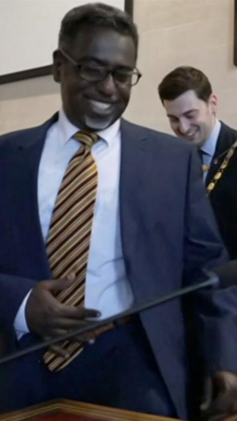 Yassin Mohamud is standing and smiling. He is wearing a blue suit, white shirt and a yellow and claret tie. He is inside a council chamber and the former Lord Mayor is in the background, he is also smiling.