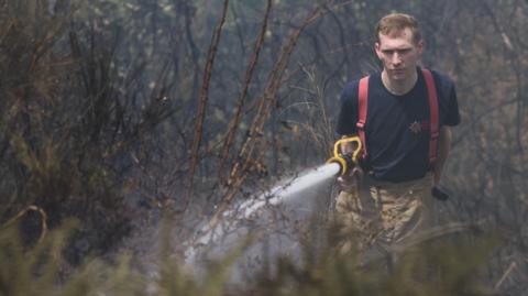 A male firefighter in a navy blue branded T-shirt, and tan brown trousers, points a hose with water coming out at dry grass.