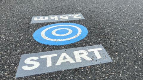 There are three parts to the marker which is on a tarmac path. The middle bit has five blue and white concentric circles, one block to the left of it reads: 2.5km and the other to the right reads: START.