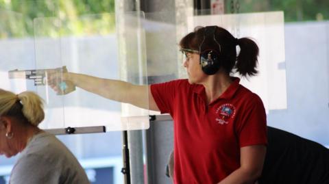 Jersey shooter Nicola Holmes, wearing a red top, points a gun