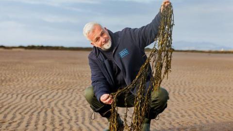 A man with grey hair and a grey beard in green trousers and wellies with a blue jumper and blue zip-up fleece holds up a long string of seaweed on a wide, sandy beach