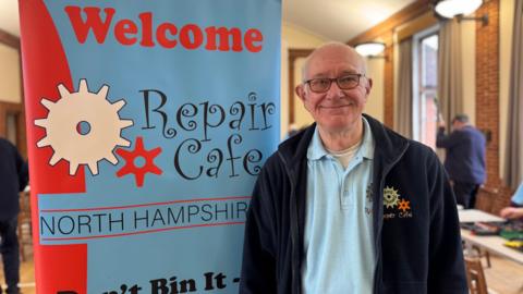 A man with white hair and glasses, wearing a navy fleece and blue polo shirt, both embroidered with a "repair cafe" logo. He is stood next to a blue and red banner which reads "welcome, repair cafe North Hampshire".