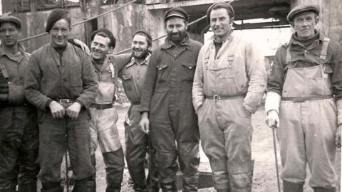 A group of seven whalers posing for the camera in a black and white picture. All are looking at the camera except the man in waders third from the left, who is staring at a man in overalls in a hat. The man on the near right is wearing waders and is leaning on a stick.