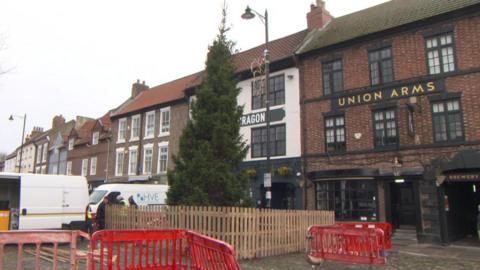 The tall, thin Christmas is outside a pub and is surrounded by a fence and there are red street barriers beside it.