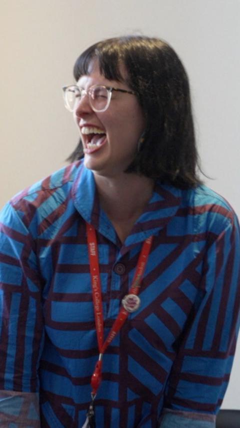 Woman with brown bob and glasses looks to her right and laughs, she is wearing a blue and red striped shirt and a red lanyard