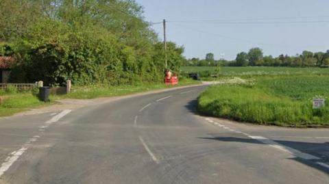 The junction of a main road with two side roads surrounded by fields and trees. 