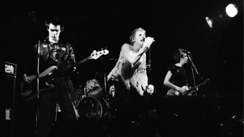 The Sex Pistols on stage in 1977. It is a black and white photo. Johnny Rotten is seen snarling into a microphone in cut up-clothing.  Sid Vicious wears a leather jacket while Steve Jones is in a crop black top. 