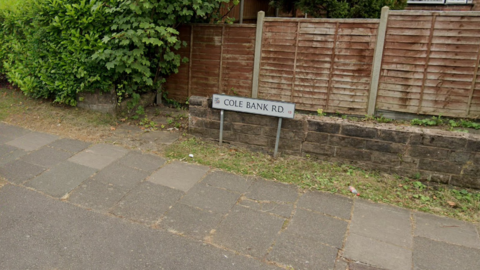 A picture of a road sign saying Cole Bank Road, in front of a very low brick wall which is three bricks high and a house fence. It is on a very thin strip of grass next to a pavement.