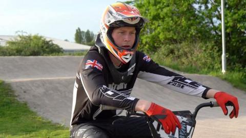 George Hunt sits on his BMX bike on a track. He is wearing cycling gear and red gloves with a helmet. There are trees around the track.