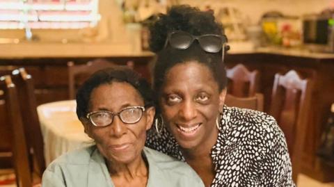 A woman with black hair in a bun, a black and white animal print top and silver hoop earrings is bend down to pose next to another woman, who is sitting on a chair. The woman on the chair has black hair which is pulled back, and black and gold glasses, with a green top.