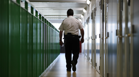 A stock photo of a male prison guard with his back to the camera, walking down a prison wing. The doors are on the right and green railings are to the left.