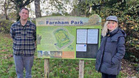 A man stood to one side of an information sign in a park and a woman stood to the right. They are both looking into the camera. Behind them is grass and trees