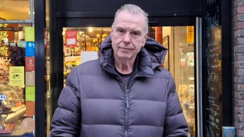 Peter Kinsella has short grey hair and is wearing a blue puffer winter coat. He is stood at the entrance to his restaurant Lunya.