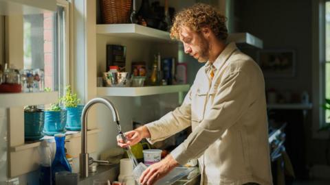 Man with curly hair wearing a beige shirt. He is washing dishes.