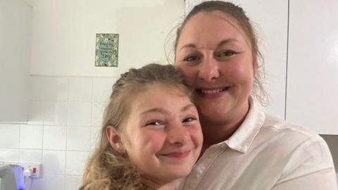 A medium close-up image of a mother and daughter standing close to each other while standing in their kitchen