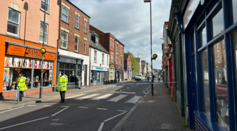 A picture shows All Saints Road, Gloucester, closed by police. Blue and white tape is visible across the street and several police officers in high-visibility jackets can be seen both in the foreground and background. 