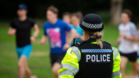 A female police officer watches park runners pass on Southampton Common. The viewer is looking at the back of her head and the runners are out of focus in the distance. She is wearing a yellow hi-viz jacket and a black vest over the top.