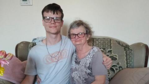 Two people sit close together on a sofa in a living room, with drinks and snacks on a table in front of them.
