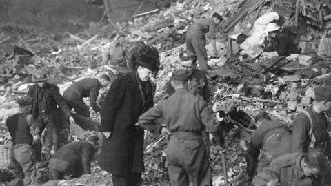 Black and white picture of men, some in uniform searching amongst rubble.
