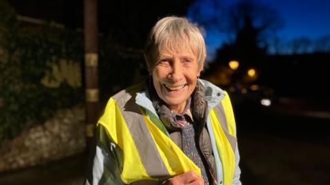 Penny Aeberhard looks at the camera and is wearing a high vis jacket. She is looking at the camera and smiling and has short grey hair.