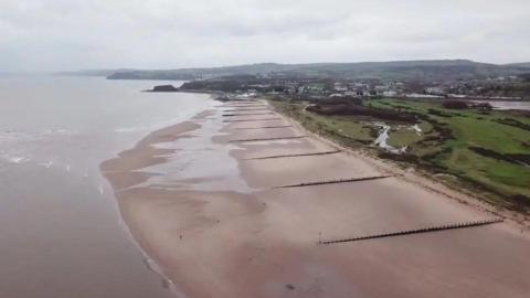 A drone shot from above Dawlish Warren beach in Devon. You can see the sea, beach, a large green area and homes and buildings in the distance. 