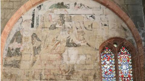 The photo shows the inside wall of a church. On the right is a small stain glass window, while to the left of it can be seen the faded detail of a painting on the wall. The outline of several figures can just about be made out.