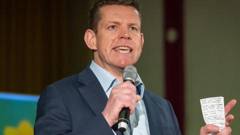 Rhun ap Iorwerth holding a set of notes while speaking into a microphone.,