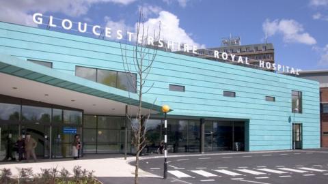 The front exterior of Gloucestershire Royal Hospital. It is a large rectangular building clad in blue panels. On the left there is a sheltered area, and a zebra crossing heading. It is a sunny day with wispy clouds in the sky.