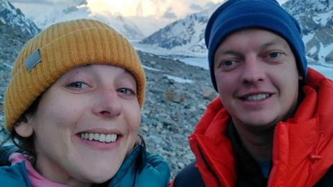 Kat Von Goldacker and Iain Bisset. They are stood together in front of a mountainous scene which looks like it is at high-altitude, given the snow, high peaks behind them and the warm mountaineering clothing they are wearing. Von Goldacker is wearing a yellow wool hat and a blue down jacket, while Bisset is wearing a navy wool hat and a red down coat.
