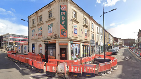 The building stands on the corner of a street with a set of roadworks surrounding the front of it. The building looks to have three levels and appears to be a shop with green and orange advertising signage on it, also filling windows. The shop, called Amber, has a vertical sign with the name on it on the corner of the building.